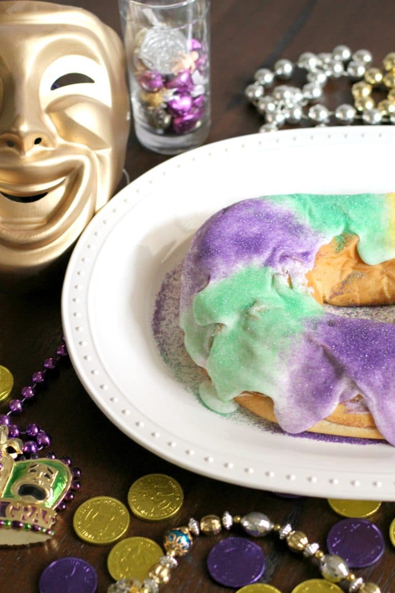 King cake on a white plate with a mardi gras mask and beads in the background. 