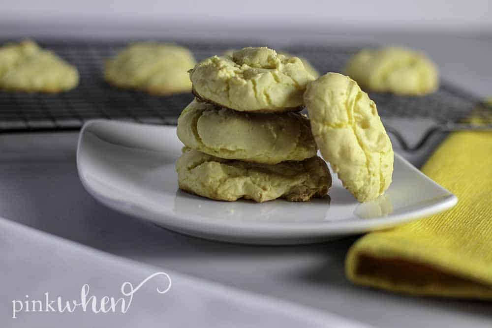 Lemon Cake Mix Cookies stacked on a white plate with cookies cooling in the background on a wire rack. 