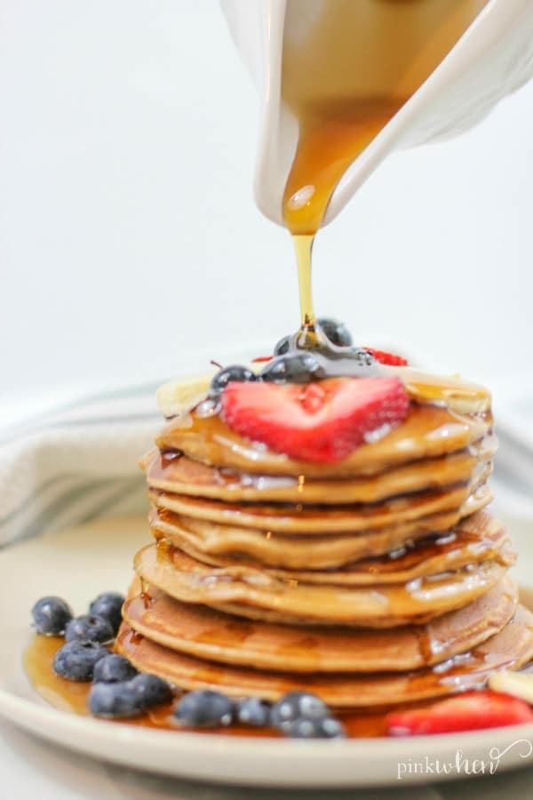 Protein pancakes covered in fruit and with syrup drizzled stacked on a white plate.
