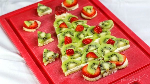 Christmas Fruit Pizza in the shape of a Christmas tree covered in cream cheese frosting and fruit on a red cutting board. 