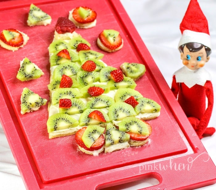 Christmas Fruit Pizza with cream cheese frosting and covered in fruit on a red cutting board with an elf looking on. 