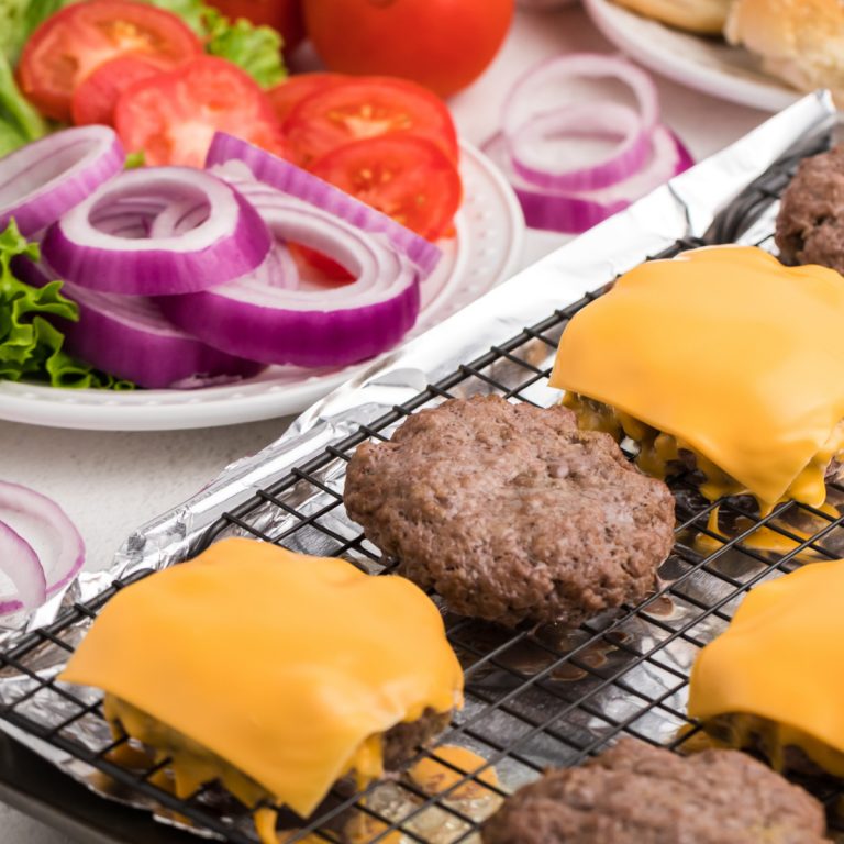 Baked hamburger patty's on a baking sheet with hamburger toppings in the background.