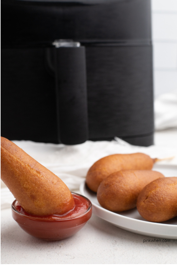 Cooked corn dog being dipped in ketchup with a plate of corn dogs and an Air Fryer basket in the background.