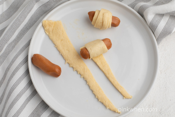 Cocktail sausages being wrapped in crescent dough on a white plate.