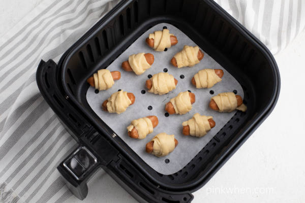 Pigs in a blanket on parchment paper in an air fryer basket ready to cook.