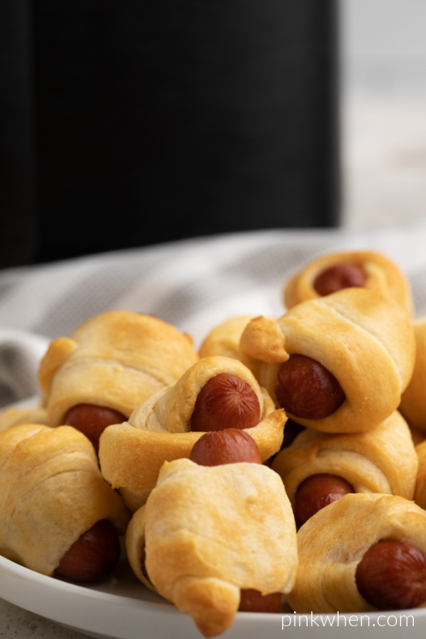 Air Fryer cooked Pigs in a Blanket on a white plate with an air fryer in the background.