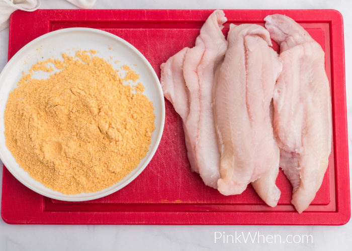 Catfish on a red cutting board and a small shallow plate with fish fry coating. 