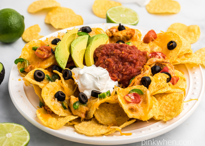 Air Fryer Nachos on a table surrounded by nachos and limes, and the nachos are piled onto a while plate. Air Fryer Nachos are also topped with sliced olives, sliced avocado, salsa, and sour cream. 