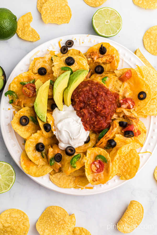 Air Fryer Nachos are layered onto a serving plate and topped with sliced olives, avocado slices, salsa, and sour cream.