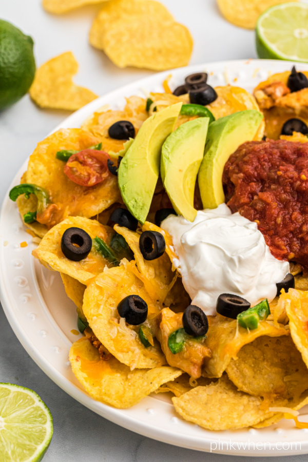 Air Fryer Nachos on a white serving plate with sliced olives, salsa, sliced avocado, and a dollop of sour cream on top. 