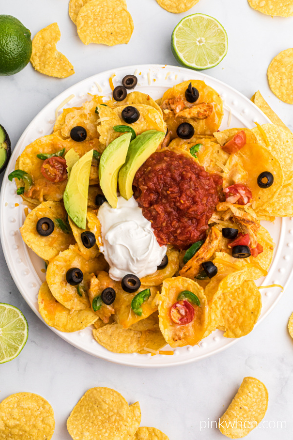 Overhead shot of Air Fryer Nachos on a white plate topped with sliced olives, avocado, salsa, and sour cream. Surrounded by chips, avocado, and limes. 