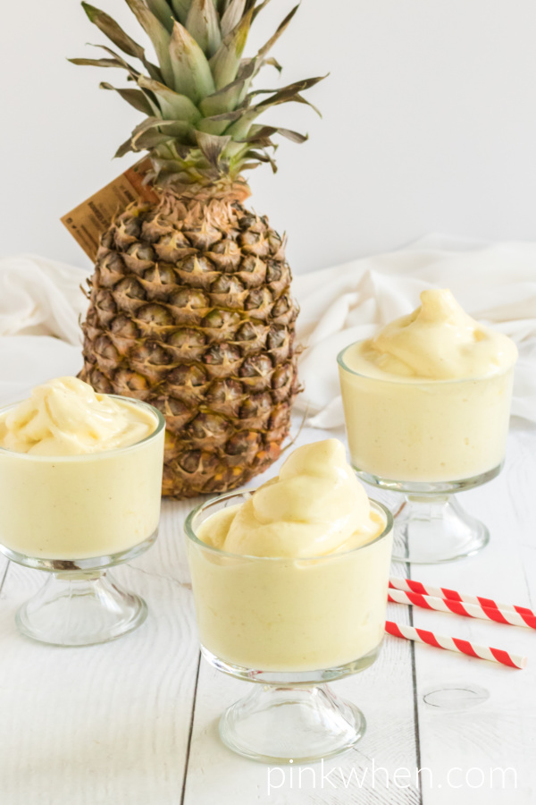 Three glasses with Dole Whip with red and white straw, pineapple in the background, and on a white table. 