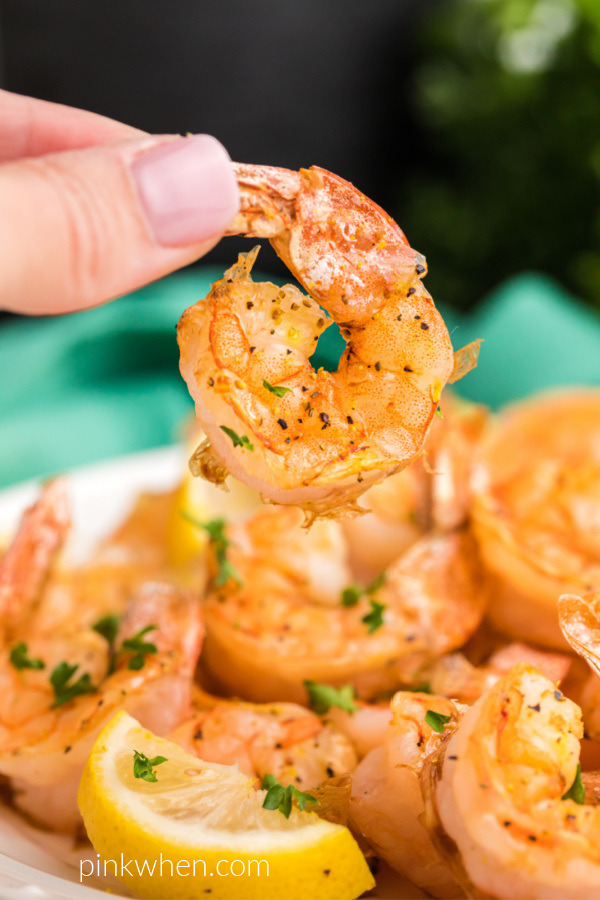 Close up photo of hand holding a lemon pepper shrimp over a plate full of shrimp and lemon wedges. 