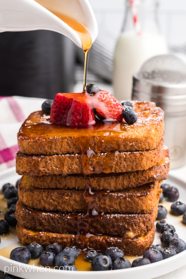 Stack of Air Fried French Toast with fresh berries on a white plate topped with confectioners' sugar and having maple syrup poured on top. 
