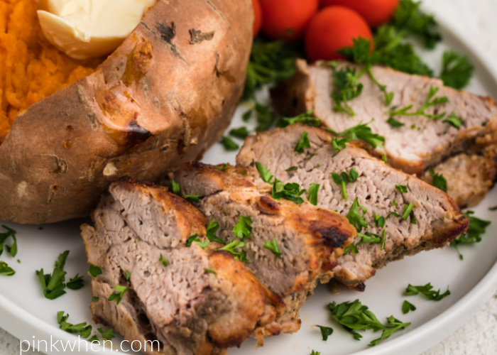 Air Fried Pork Tenderloin on a ehite plate with a sweet potato and tomatoes and garnished with fresh parsley. 