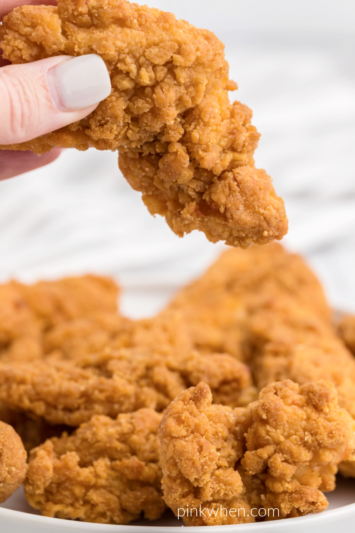 Fully cooked frozen air fryer chicken strips on a white plate with a hand holding a chicken strip overhead.