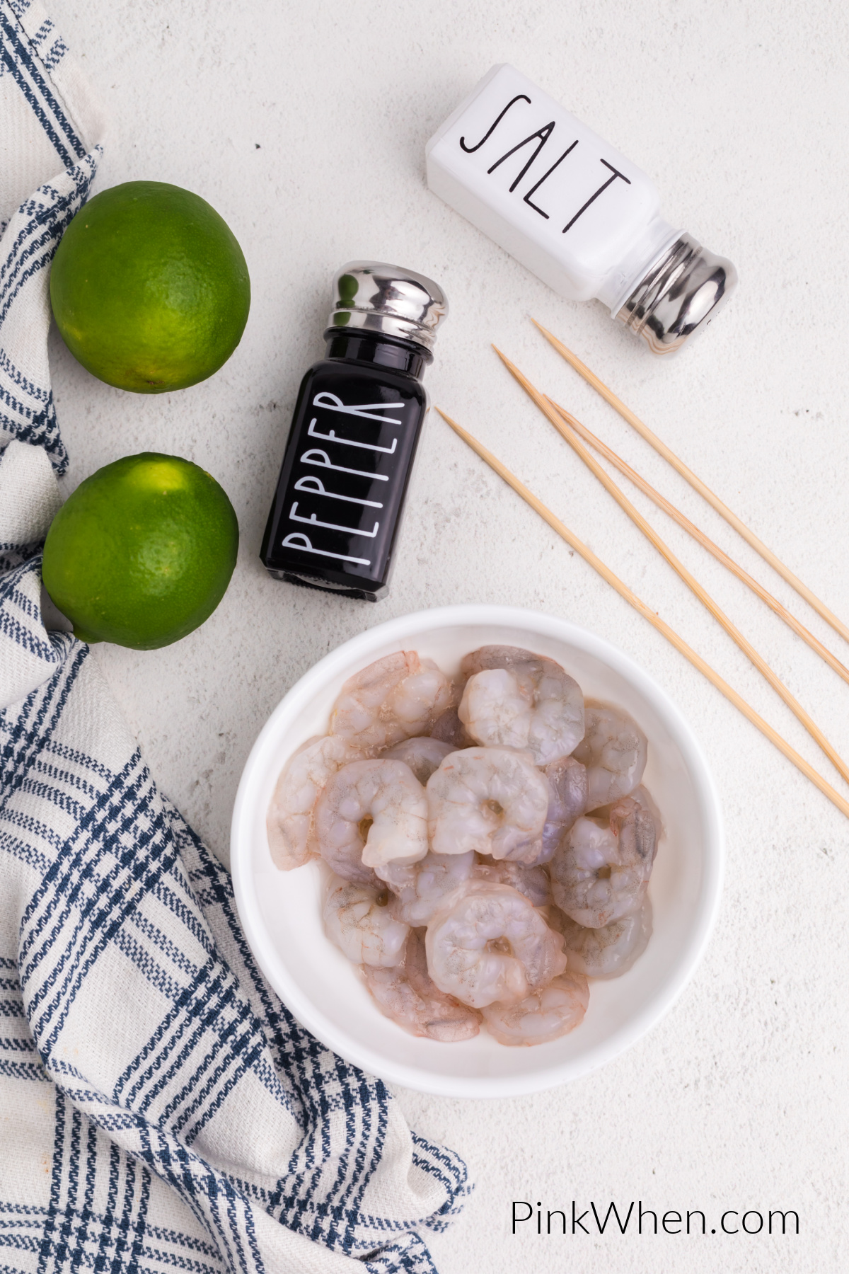 Bowl full of fresh raw shrimp, wooden skewers, limes, salt, and pepper.