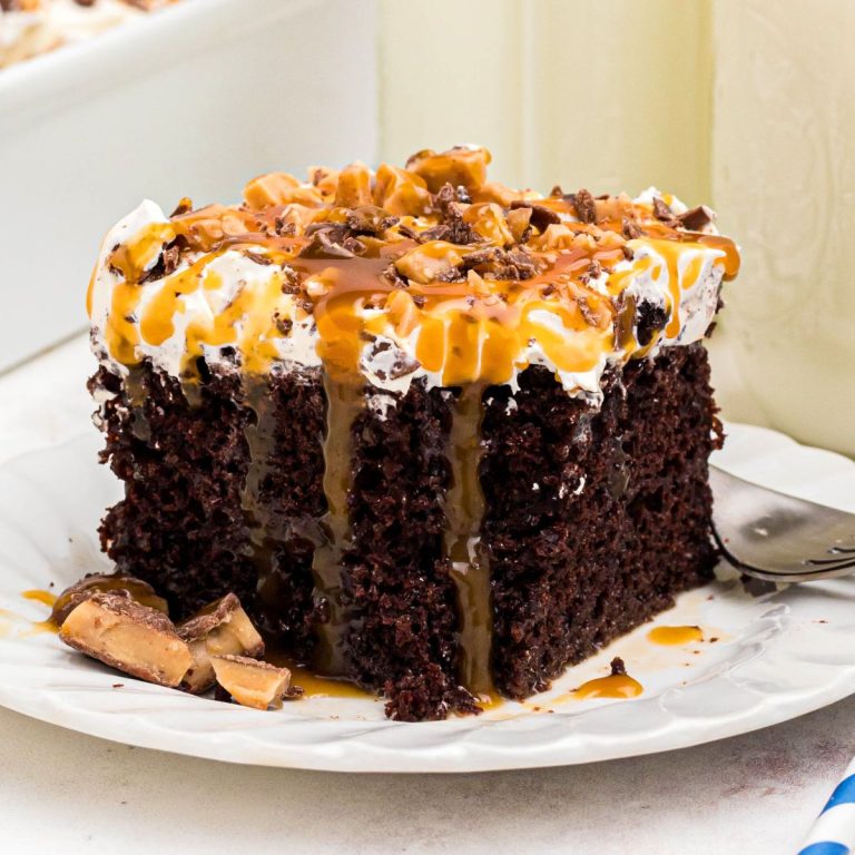 Chocolate slice of cake on a white plate, topped with whipped cream, candy bits, and caramel sauce.