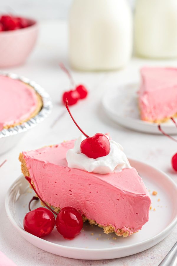 Pink slice of kool aid pie on a small white plate with cherries for a garnish