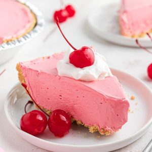 Pink slice of cherry kool aid pie on a small white plate topped with cool whip and cherries