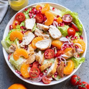 Leafy greens tossed with orange slices, vegetables, and chicken pieces in a white bowl.