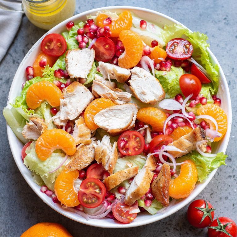 Leafy greens tossed with orange slices, vegetables, and chicken pieces in a white bowl.