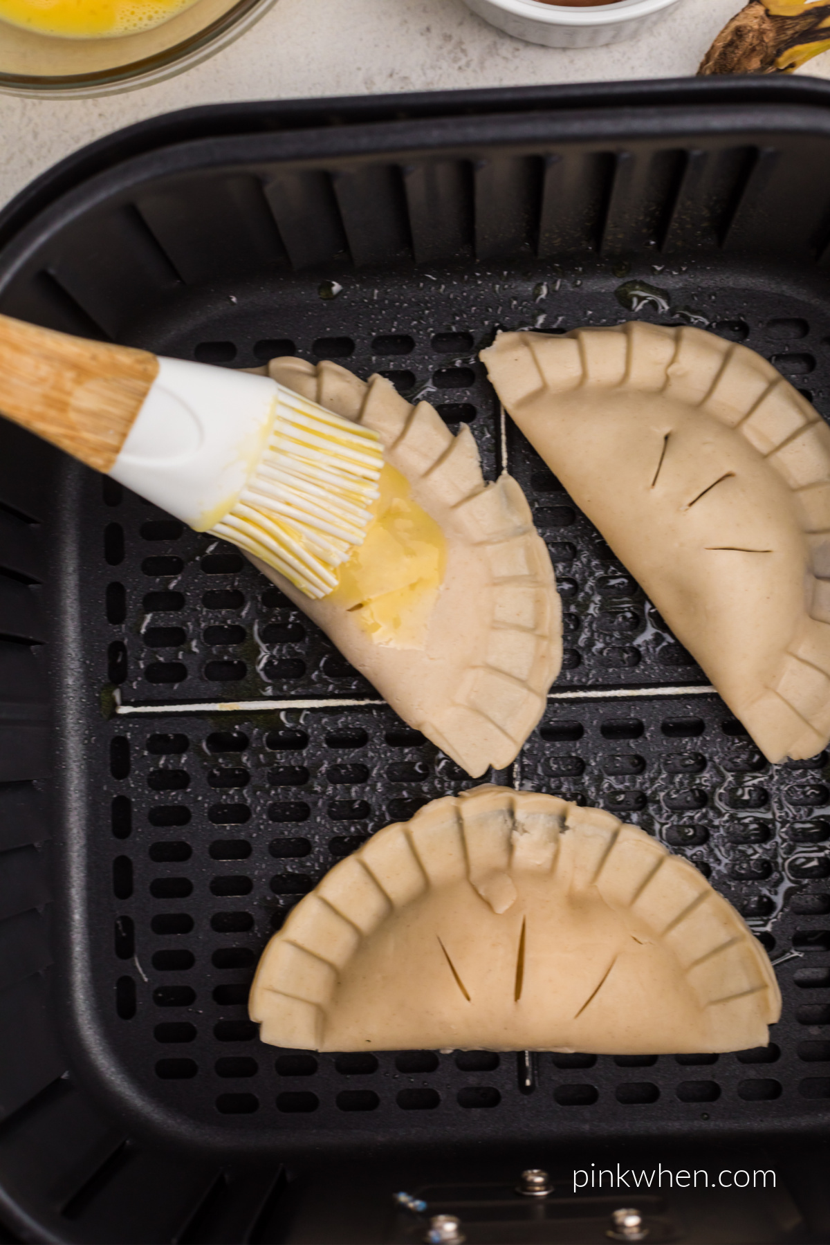 Mini hand pies in the basket of the air fryer being brushed with egg mixture before cooking.