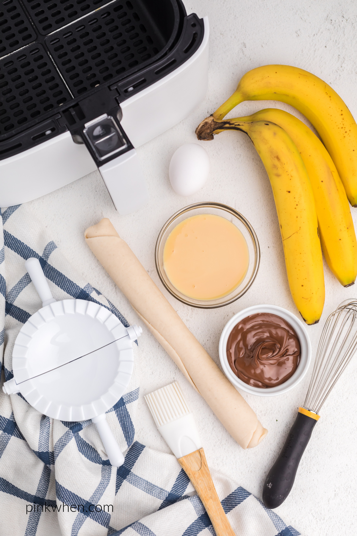 Nutella, bananas, sweetened condensed milk, egg, brush, and pie cutter on a white table. 