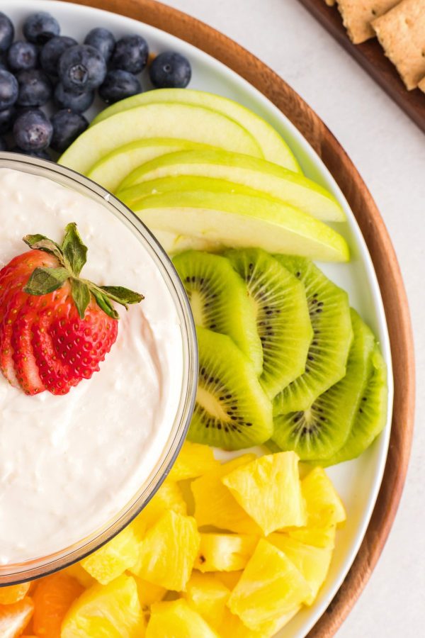 Bright fresh fruits on a white platter with a bowl of creamy fruit dip in the center of the platter. 