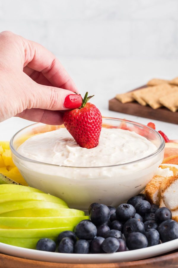 Bright red strawberry being dipped into fruit dip in a clear glass bowl surrounded by other fruit. 