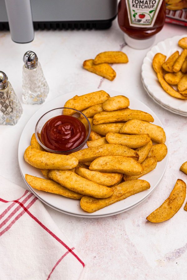 Golden steak fries on a white plate with ketchup in front of an air fryer. 