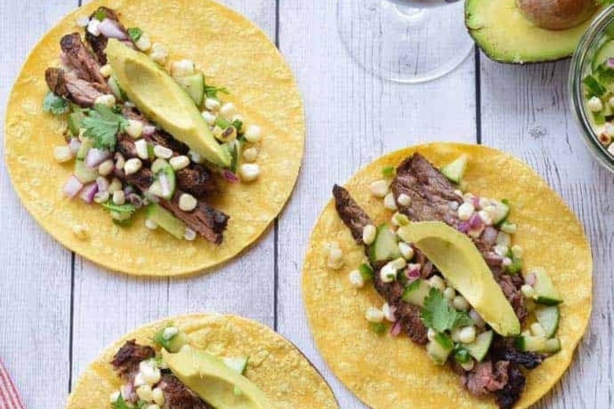 Three tacos with steak and corn salsa on a wooden table.