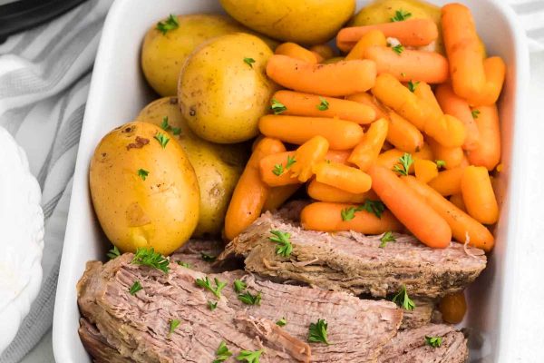 Slow cooker beef roast with carrots and potatoes in a white dish.