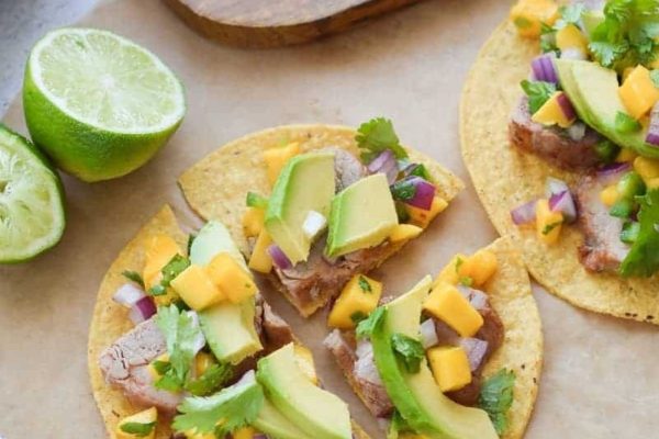 Meat tacos with mango salsa and lime wedges on a cutting board.