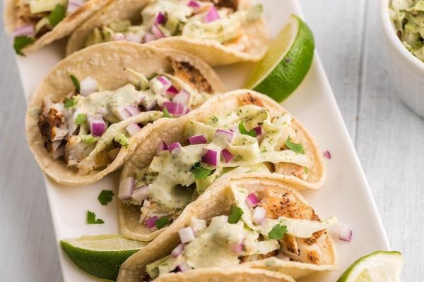 Four tacos with guacamole and lime on a white plate.