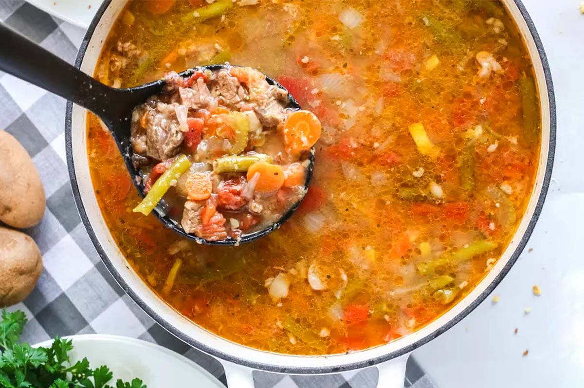 A ladle scooping vegetable beef soup from a pot.
