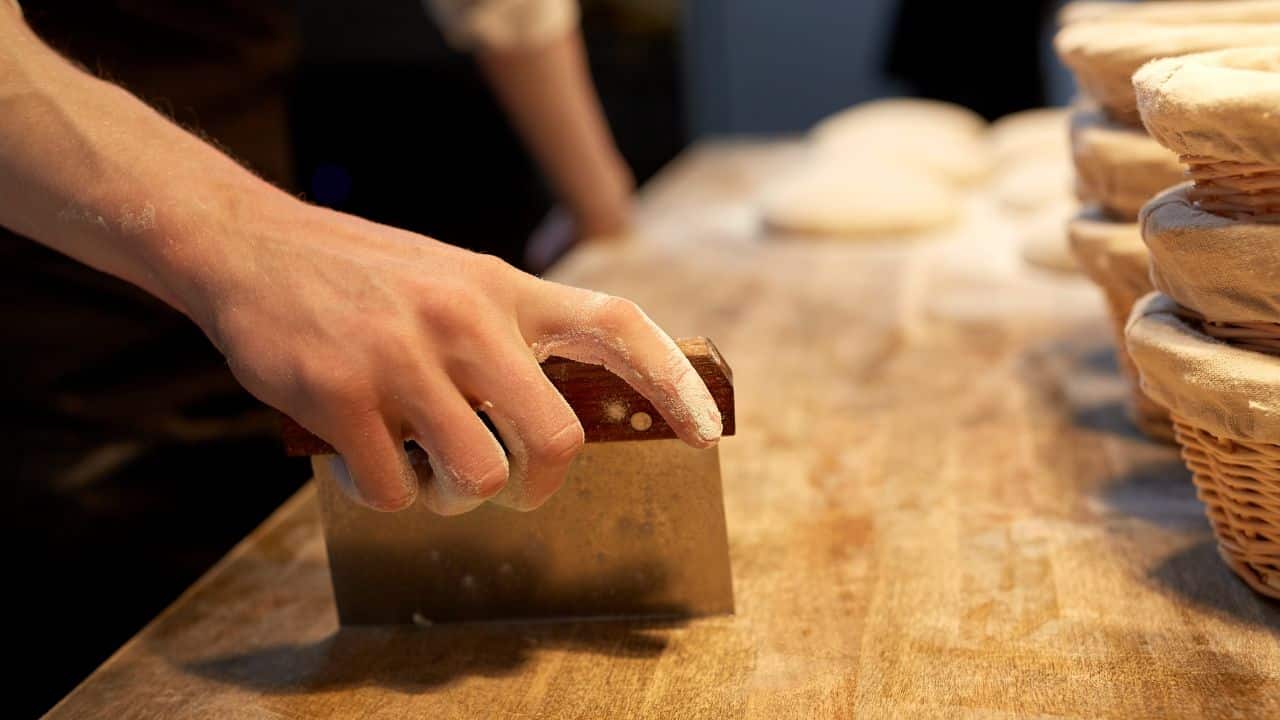 Hand holding a bench scraper on a wooden chopping block.