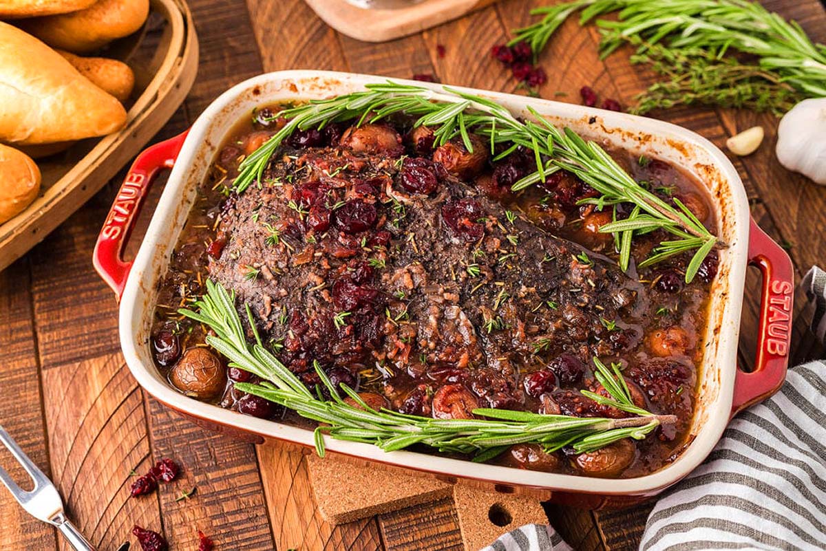 A brisket in a casserole dish with roasted cranberries and fresh herbs.