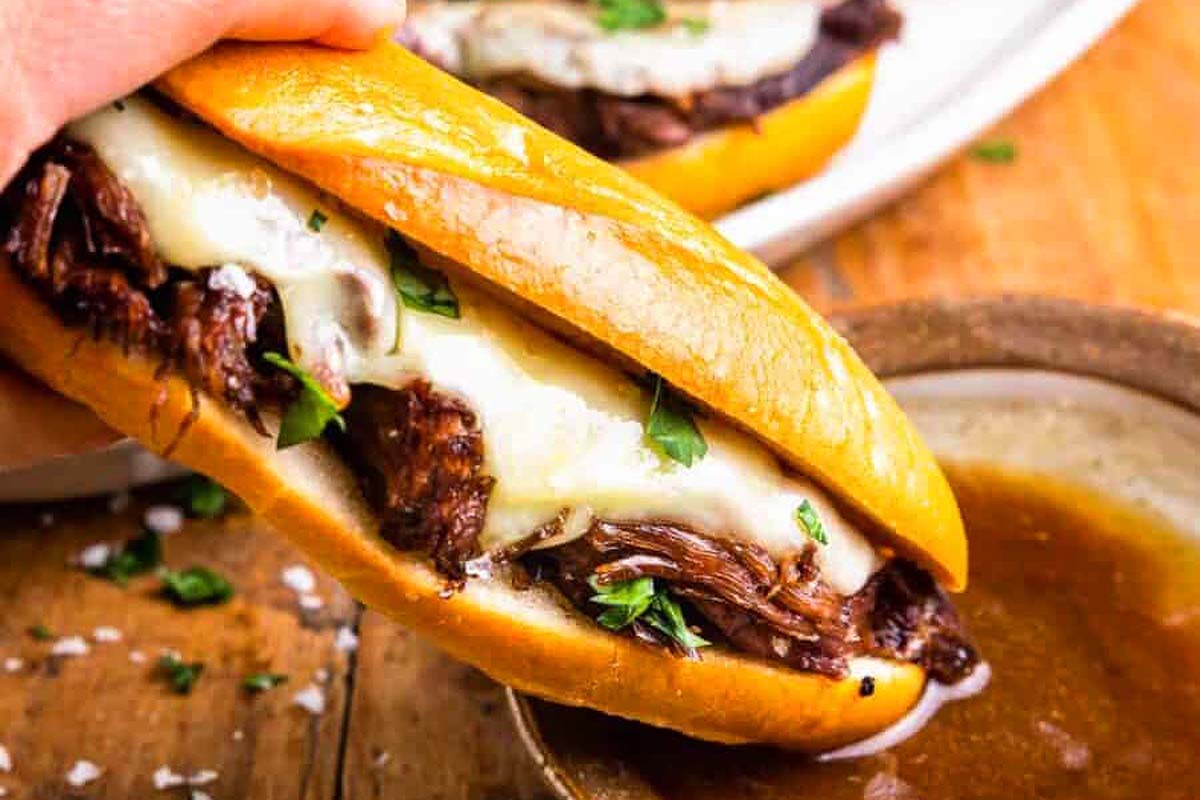 A hand dipping the french dip sandwich into the au jus.
