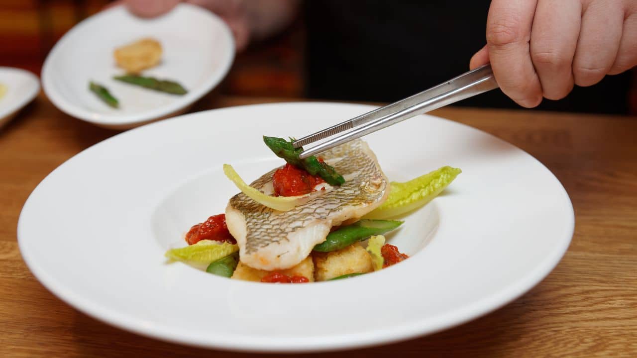 Chef is decorating a fish dish with asparagus using kitchen tweezers.