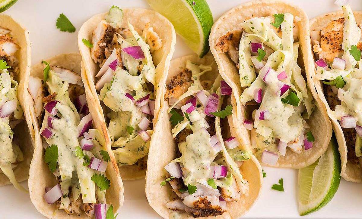 Four fish tacos on a white plate with lime wedges.