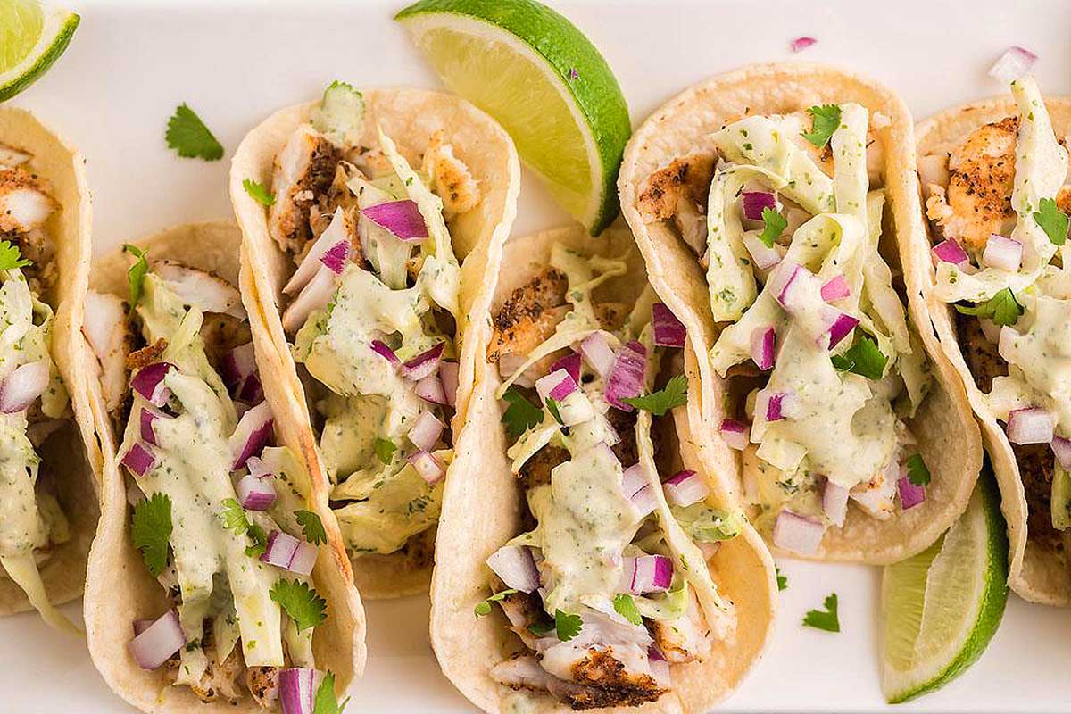 Four fish tacos on a white plate with lime wedges.