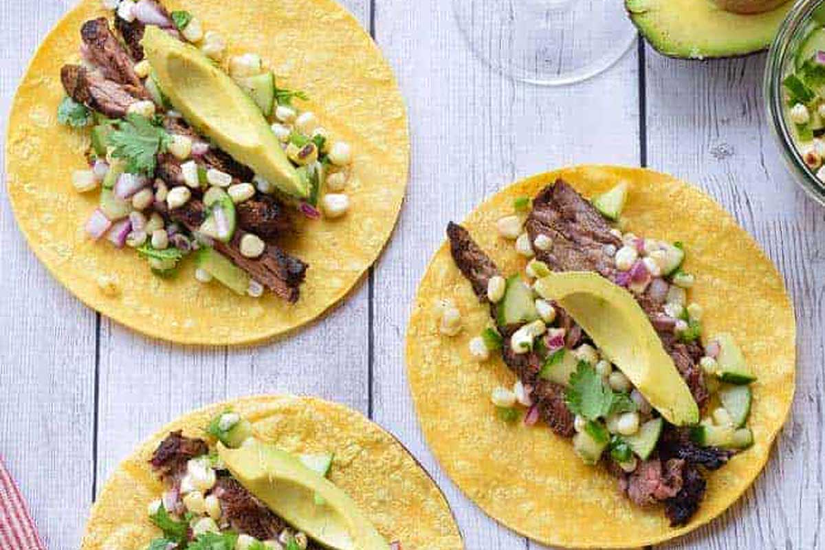 Three tacos on a wooden table with avocado and corn.