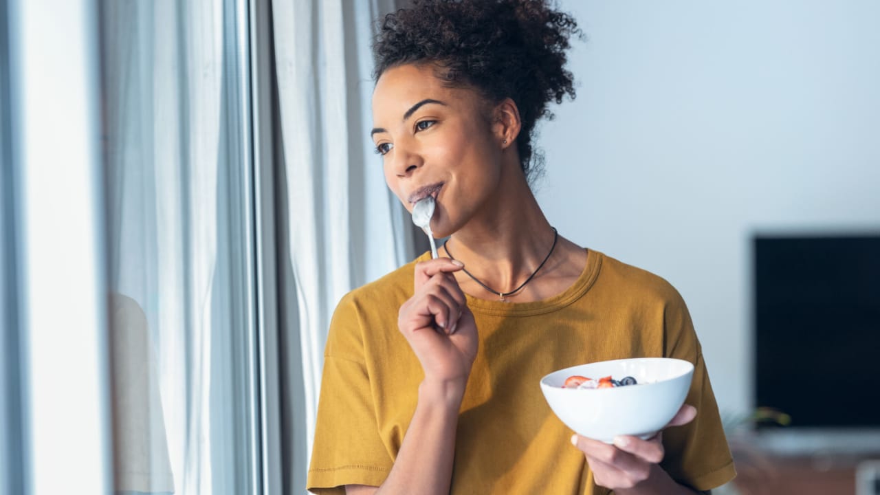 Beautiful woman eating yogurt while standing next to the window at home.