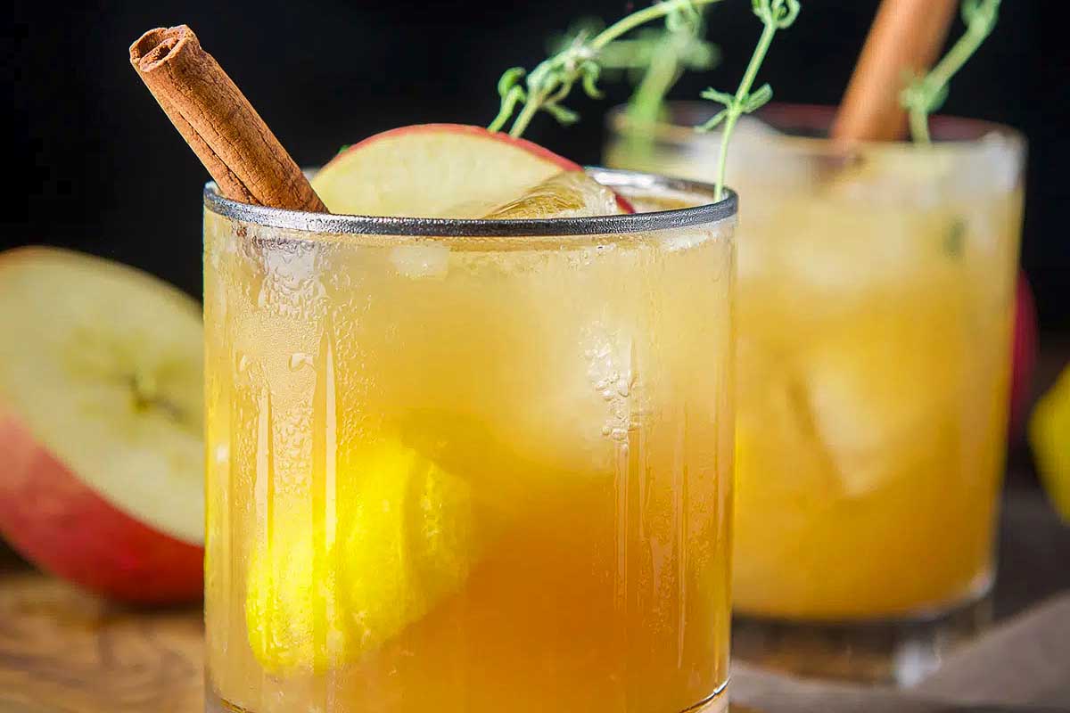 Two glasses of apple cider with cinnamon sticks and apples, perfect for Fall gatherings.