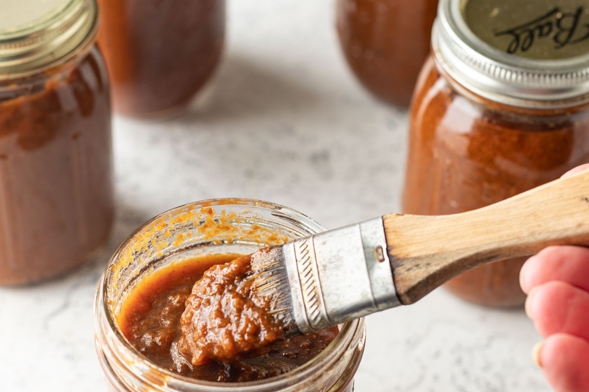 A person is dipping a brush into a jar of sauce for their recipe.