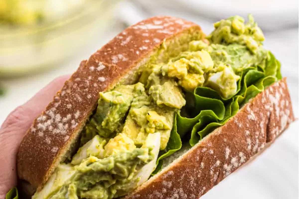 A person holding a sandwich with guacamole and lettuce.