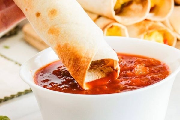 A person is dipping a burrito into a bowl of sauce during their meal prep.