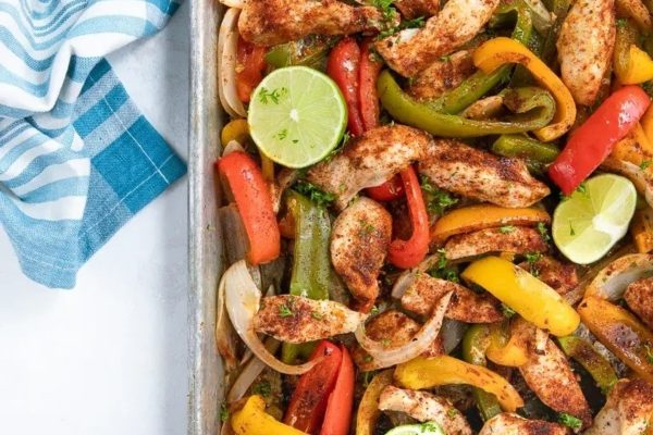 Chicken fajitas on a baking sheet with peppers and lime wedges.