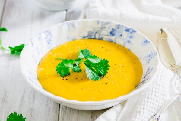 A bowl of carrot soup with parsley on a wooden table with few ingredients.